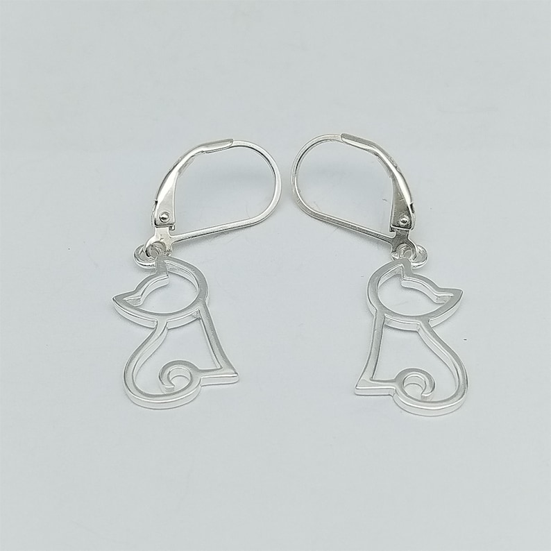May include: A pair of silver dangle earrings featuring a simple outline of a cat. The earrings have a leverback closure.