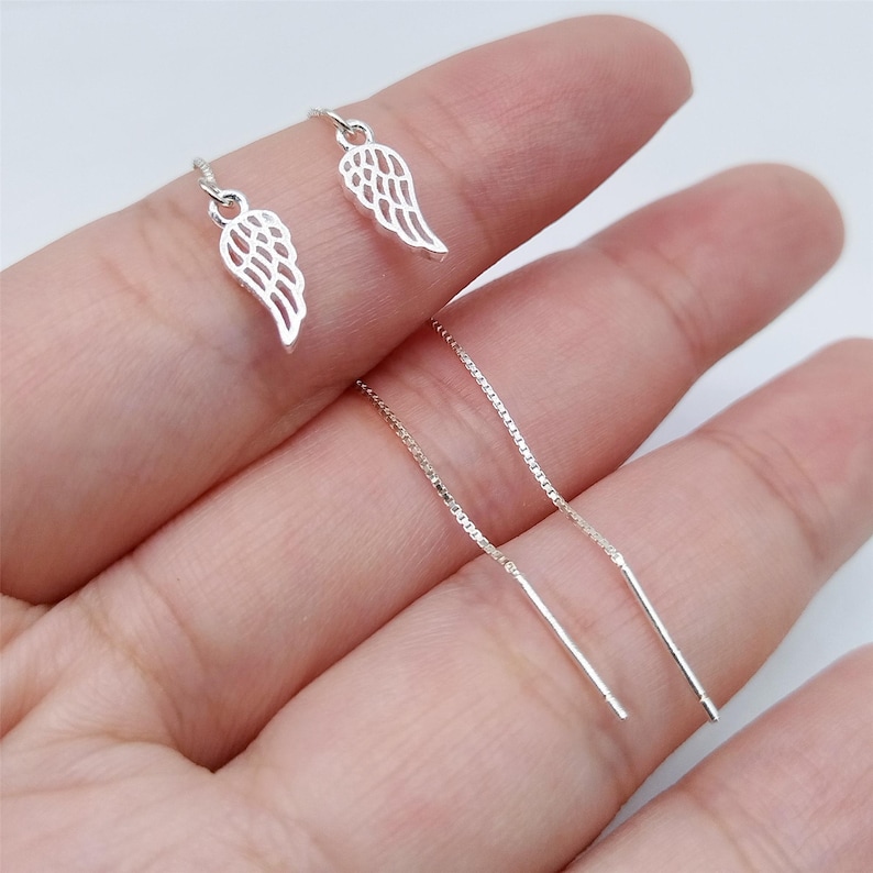 May include: Silver-coloured dangle earrings with a wing design. The earrings have a delicate, openwork pattern and are suspended from a fine chain. The earrings are displayed on a person's finger.