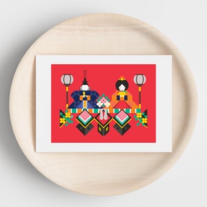 May include: A red card with a white border features a stylized illustration of two dolls in traditional Japanese clothing. The dolls are surrounded by colorful geometric shapes and flowers. The card is on a wooden surface.