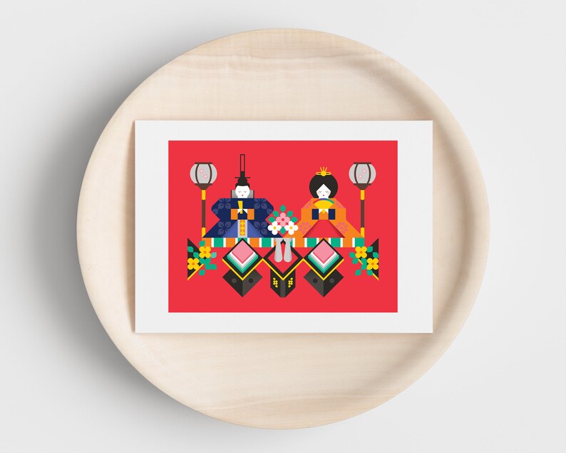 May include: A red card with a white border features a stylized illustration of two dolls in traditional Japanese clothing. The dolls are surrounded by colorful geometric shapes and flowers. The card is on a wooden surface.