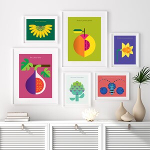 May include: A set of nine framed prints featuring colorful illustrations of various fruits, vegetables, and insects. The prints are arranged in a grid pattern on a white wall above a white cabinet with two doors.