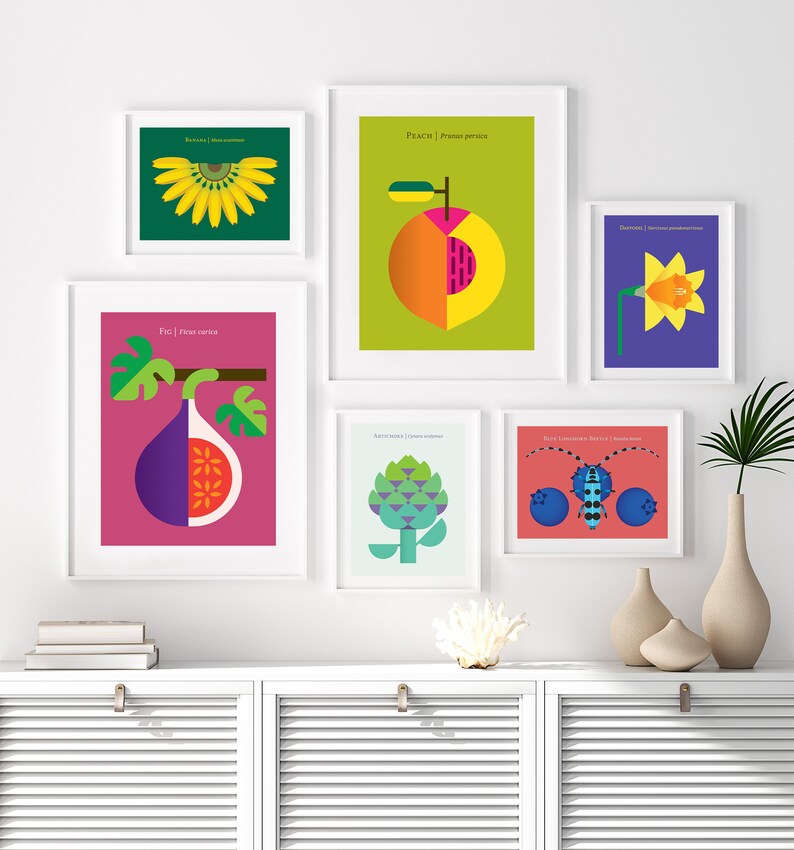May include: A set of nine framed prints featuring colorful illustrations of various fruits, vegetables, and insects. The prints are arranged in a grid pattern on a white wall above a white cabinet with two doors.