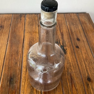 May include: A clear glass bottle with a black cap and cork stopper. The bottle has a rounded base and a long neck. The bottle is sitting on a wooden surface.