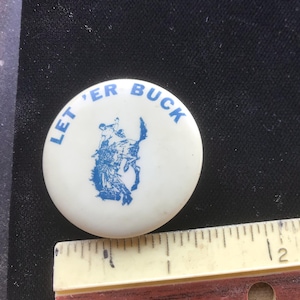 May include: A white button with blue lettering that reads "Let 'er Buck". A blue silhouette of a cowboy riding a bucking bronco is in the center of the button.