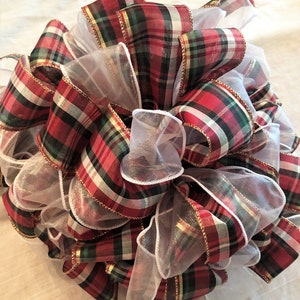 May include: A decorative bow made of red, green, and gold plaid ribbon with white sheer ribbon accents. The bow is tightly wound and has a full, round shape, perfect for holiday decorations.