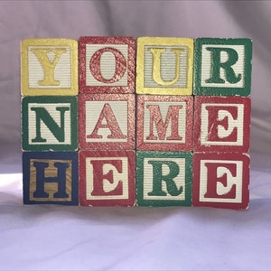 May include: A set of twelve square wooden blocks arranged in a three-by-four grid. The blocks feature alternating red, green, yellow, and blue colors with embossed letters spelling out the phrase YOUR NAME HERE in a mix of gold, red, and green.