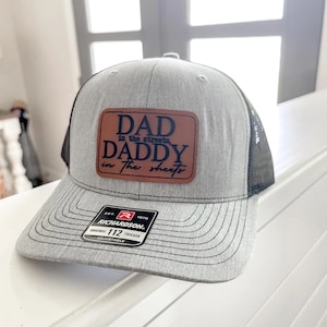 May include: A gray and black trucker hat with a brown leather patch that says "DAD in the streets, DADDY in the sheets". The hat is adjustable and has a Richardson logo.