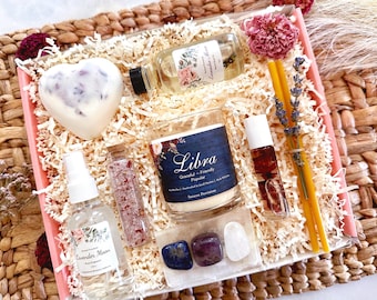 May include: A pink gift box filled with items for a Libra zodiac sign. The box contains a candle, bath salts, a heart-shaped bath bomb, a bottle of essential oil, a roll-on essential oil, and two crystals. The candle has a blue label with the word "Libra" and the text "Gracious - Friendly - Popular".