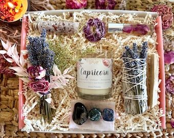 Op de afbeelding: Een cadeaudoos gevuld met items voor het sterrenbeeld Steenbok. De doos bevat een kaars, een salie-reinigingsstok, een fles badzout, kristallen en gedroogde bloemen. De kaars is gelabeld "Steenbok: Verantwoordelijk - Geduldig - Volhardend".