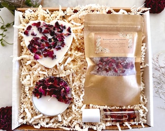 May include: A gift box filled with bath and body products. The box contains a heart-shaped candle with dried rose petals, a small bag of bath salts with rose petals, and a bottle of essential oil. The box is filled with shredded paper.