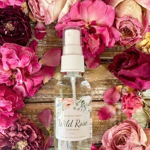 May include: A clear glass bottle with a white spray nozzle and a label that reads "Wild Rose Room Fragrance" by Eternal Zen Creations. The bottle is surrounded by dried pink rose petals.