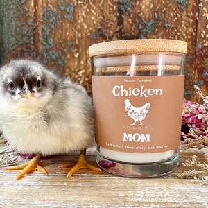 May include: A brown glass candle jar with a bamboo lid. The label reads "Chicken" with a white chicken illustration and the text "MOM" below. The candle is soy wax, handcrafted, and made with love. A small, fluffy, gray and white chick is standing in front of the candle.