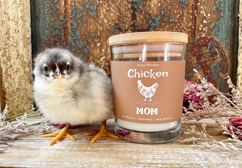 May include: A brown glass candle jar with a bamboo lid. The label reads "Chicken" with a white chicken illustration and the text "MOM" below. The candle is soy wax, handcrafted, and made with love. A small, fluffy, gray and white chick is standing in front of the candle.