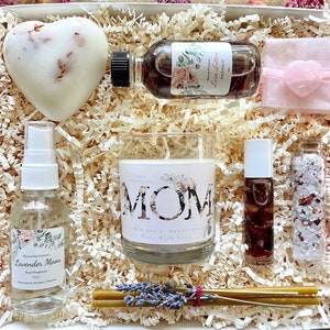 May include: A gift box filled with self-care items for Mother's Day. The box includes a white heart-shaped bath bomb, a candle with the word "MOM" on it, a bottle of lavender moon room fragrance, a bottle of rose-scented bath oil, a bottle of rose-scented bath salts, and a small pink heart-shaped soap.
