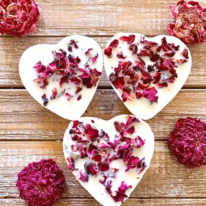 Pu&ograve; includere: Tre saponi bianchi a forma di cuore con petali di rosa secchi incorporati. I saponi sono disposti su una superficie di legno con boccioli di rosa secchi sparsi intorno.
