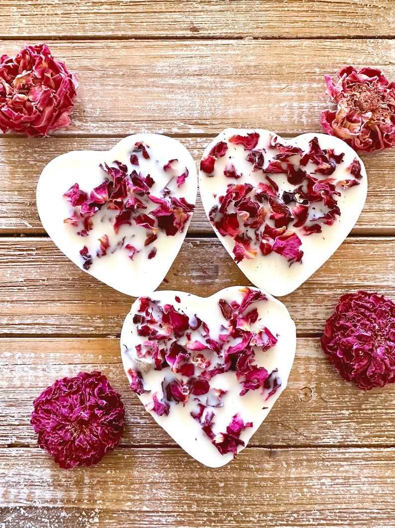 Pu&ograve; includere: Tre saponi bianchi a forma di cuore con petali di rosa secchi incorporati. I saponi sono disposti su una superficie di legno con boccioli di rosa secchi sparsi intorno.