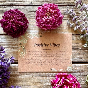 Pu&ograve; includere: Un biglietto marrone con il testo "Positive Vibes" e "Orange Calcite" su di esso. Il biglietto &egrave; circondato da fiori secchi, tra cui lavanda e rose.