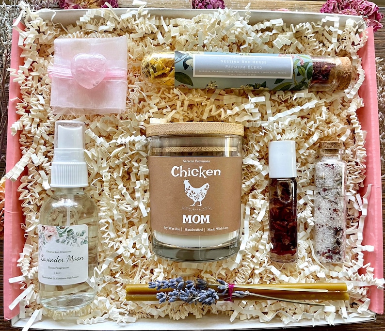 May include: A gift box filled with a variety of items, including a candle, a bottle of room spray, a small bottle of bath salts, a tube of essential oil, and a small pink heart-shaped object. The candle is labeled "Chicken Mom" and the room spray is labeled "Lavender Moon".