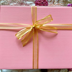 May include: A pink gift box with a gold ribbon and bow. The box is tied with a sheer gold ribbon.