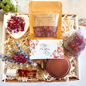May include: A gift box filled with bath and body products for Mother's Day. The box includes a heart-shaped soap, bath salts, a small bag of dried rose petals, a candle, and a bottle of essential oil. The box is decorated with a card that says "Happy Mother's Day".