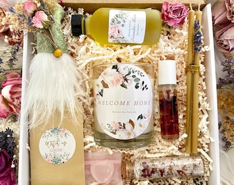 May include: A gift box filled with a variety of items, including a candle, bath salts, a gnome, and a bottle of essential oil. The candle has a floral design and the text "Welcome Home" on the label. The bath salts are in a small glass jar with a pink lid. The gnome is wearing a green hat and has a white beard. The essential oil is in a clear glass bottle with a yellow label that says "Lavender Blossom".