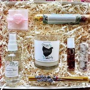 May include: A gift box filled with a variety of items, including a candle, a bottle of essential oil, a bottle of bath salts, a small bag of dried herbs, and a small piece of rose quartz. The candle is labeled "Crazy Chicken Lady" and the essential oil is labeled "Lavender Moon". The box is filled with shredded paper.
