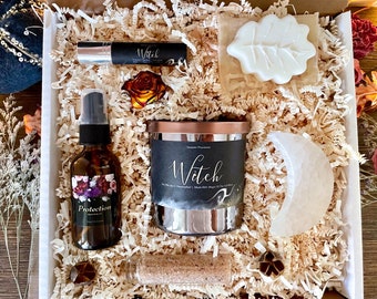 May include: A gift box filled with witchy themed items, including a candle, a soap, a spray bottle, a crystal, and a small bottle of essential oil. The candle is labeled "Witch" and the spray bottle is labeled "Protection".