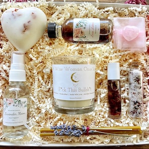 May include: A gift box filled with self-care items, including a heart-shaped bath bomb, a candle with the text "A Wise Woman Once Said F*ck This Bullsh't", a bottle of lavender moon spray, a bottle of rose oil, a small bag with a pink heart-shaped crystal, and a bottle of rose bath salts.
