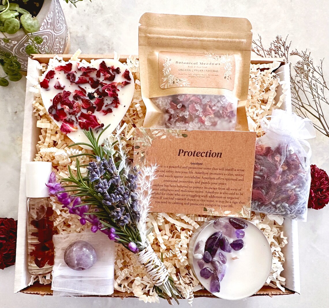 May include: A gift box filled with items for protection and spiritual cleansing. The box includes a white heart-shaped candle with dried rose petals, a small bag of dried herbs, a white candle with amethyst crystals, a small bottle of essential oil, a bundle of dried herbs tied with twine, and a card with the text "Protection" and "Amethyst".