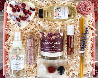 May include: A gift box filled with items for a Scorpio zodiac sign. The box includes a candle, bath salts, a spray bottle, a roll-on bottle, incense sticks, and crystals. The candle has a floral design and the text "Scorpio" on the label. The box is filled with shredded paper and has a pink lid.
