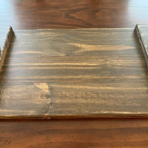 May include: A rectangular wooden serving tray with a dark brown stained finish and visible wood grain. The tray features two black metal handles attached to raised wooden ends on the left and right sides of the flat central surface.