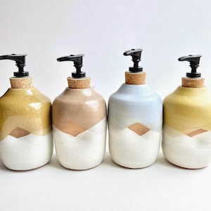 May include: Four ceramic soap dispensers arranged in a row. Each bottle features a two-tone glazed finish with a white base and a colored top in mustard, beige, light blue, or yellow. Each dispenser has a cork stopper and a black plastic pump.