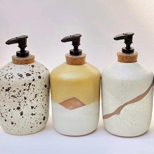 May include: Three ceramic soap dispensers with black plastic pump tops and cork stoppers. The first is cream with black speckles, the second is yellow and white with a geometric design, and the third is cream with a single brown diagonal line.