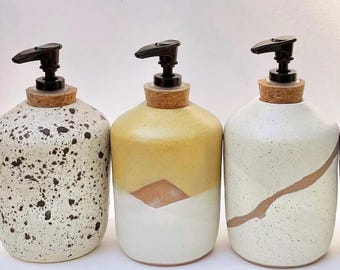 May include: Three ceramic soap dispensers with black plastic pump tops and cork stoppers. The first is cream with black speckles, the second is yellow and white with a geometric design, and the third is cream with a single brown diagonal line.