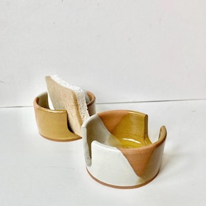 May include: Two ceramic sponge holders in the image. One is a solid mustard yellow, and the other has a white, brown, and yellow color block design. A beige sponge sits in the mustard yellow holder.