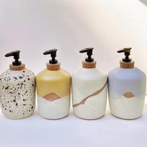 May include: Four ceramic soap dispensers with black plastic pump heads and cork stoppers. The first is white with black speckles, the second is yellow and white, the third is white with a brown diagonal line, and the fourth is light blue and white.
