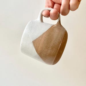 May include: A handmade ceramic mug with a geometric design. The mug features a white and brown color scheme, with a diagonal color block pattern. The handle is a rounded square shape. The mug is held by a hand against a white background.