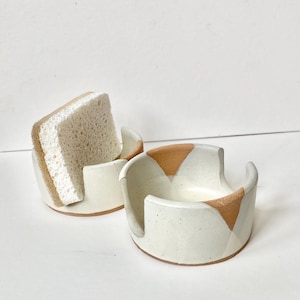 May include: Two off-white ceramic sponge holders with a speckled finish and brown accents. One holder contains a beige and brown sponge. The holders have a curved design with a cutout for the sponge.