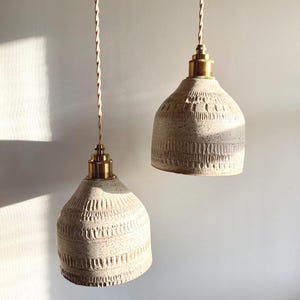 May include: Two off-white ceramic pendant lights with textured patterns and brass-colored hardware. The lights hang from twisted beige cords against a white wall. The lamps have a bell-like shape and are illuminated by sunlight.