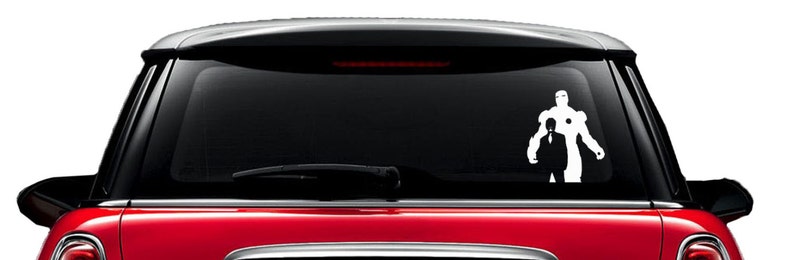 Puede incluir: Calcoman&iacute;a de vinilo blanco de un superh&eacute;roe y un hombre con traje, aplicada en la ventana trasera de un coche rojo. El superh&eacute;roe lleva un traje rob&oacute;tico, y el hombre est&aacute; a su lado.