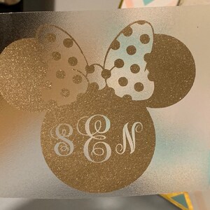 May include: A gold glitter vinyl decal featuring a stylized mouse head with a polka dot bow. The mouse head has the letters S, E, and N in a decorative font. The decal is on a clear backing.