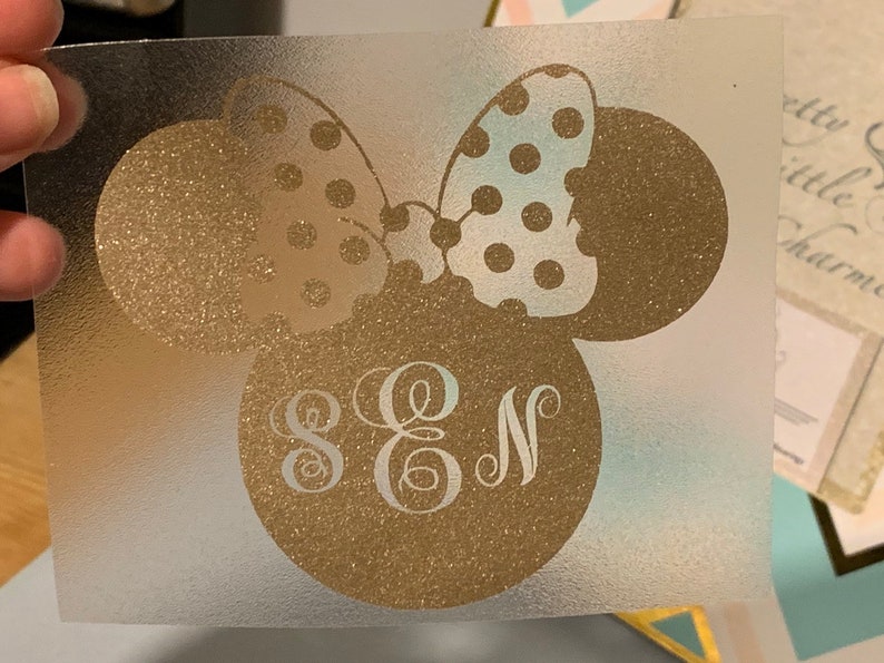 May include: A gold glitter vinyl decal featuring a stylized mouse head with a polka dot bow. The mouse head has the letters S, E, and N in a decorative font. The decal is on a clear backing.
