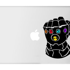 May include: Black gauntlet with six colorful gems, a design inspired by a popular superhero franchise. The gauntlet is a bold graphic on a light gray surface, likely a laptop. The gems are orange, red, blue, purple, yellow, and green.