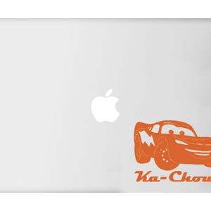 May include: Orange cartoon car sticker with the text "Ka-Chow!" applied to a white laptop. The car has large wheels and a smiling face. The sticker is a fun and playful design.