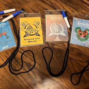 May include: Four clear plastic badge holders with black lanyards. Each badge has a different design with the words "Drinking Around The World". One badge has wine glasses, another has a yellow background, a third has a brown background, and the last has a Christmas theme.