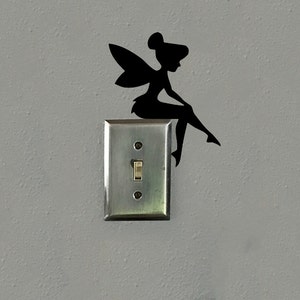 May include: Black silhouette of a fairy sitting on a light switch. The fairy has wings and is perched on the edge of the switch plate. The light switch is in the on position, and the wall is painted a light gray.