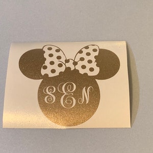 May include: A gold-colored decal featuring a Minnie Mouse head silhouette with a white polka-dot bow. The head includes the initials S, E, and N in a decorative font. The decal is on a white background.