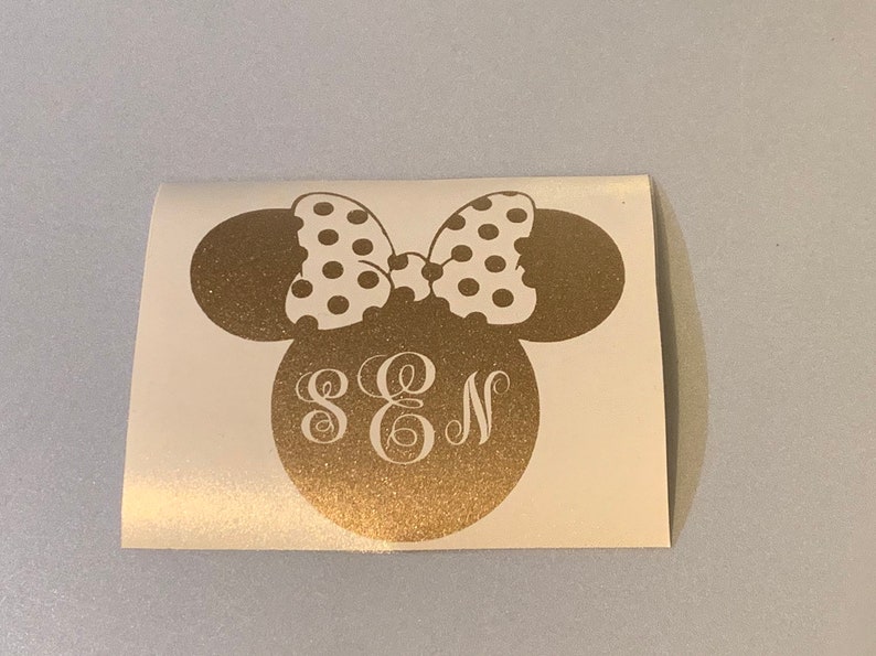 May include: A gold-colored decal featuring a Minnie Mouse head silhouette with a white polka-dot bow. The head includes the initials S, E, and N in a decorative font. The decal is on a white background.