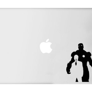 Puede incluir: Un port&aacute;til blanco con una silueta negra de Iron Man y Tony Stark. La figura de Iron Man est&aacute; a la derecha, con Stark con traje y corbata delante. El logotipo de Apple es visible en el port&aacute;til.