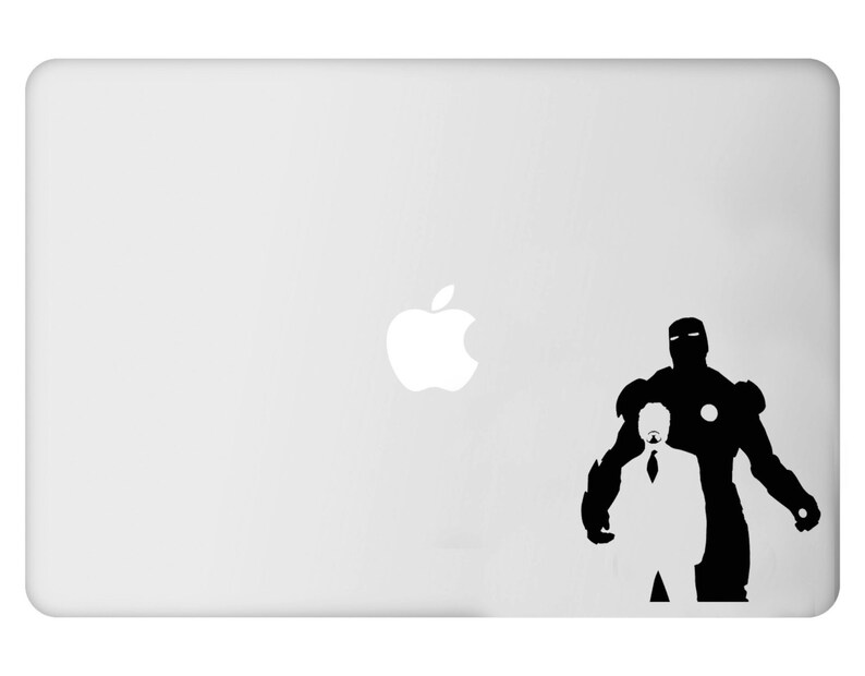 Puede incluir: Un port&aacute;til blanco con una silueta negra de Iron Man y Tony Stark. La figura de Iron Man est&aacute; a la derecha, con Stark con traje y corbata delante. El logotipo de Apple es visible en el port&aacute;til.
